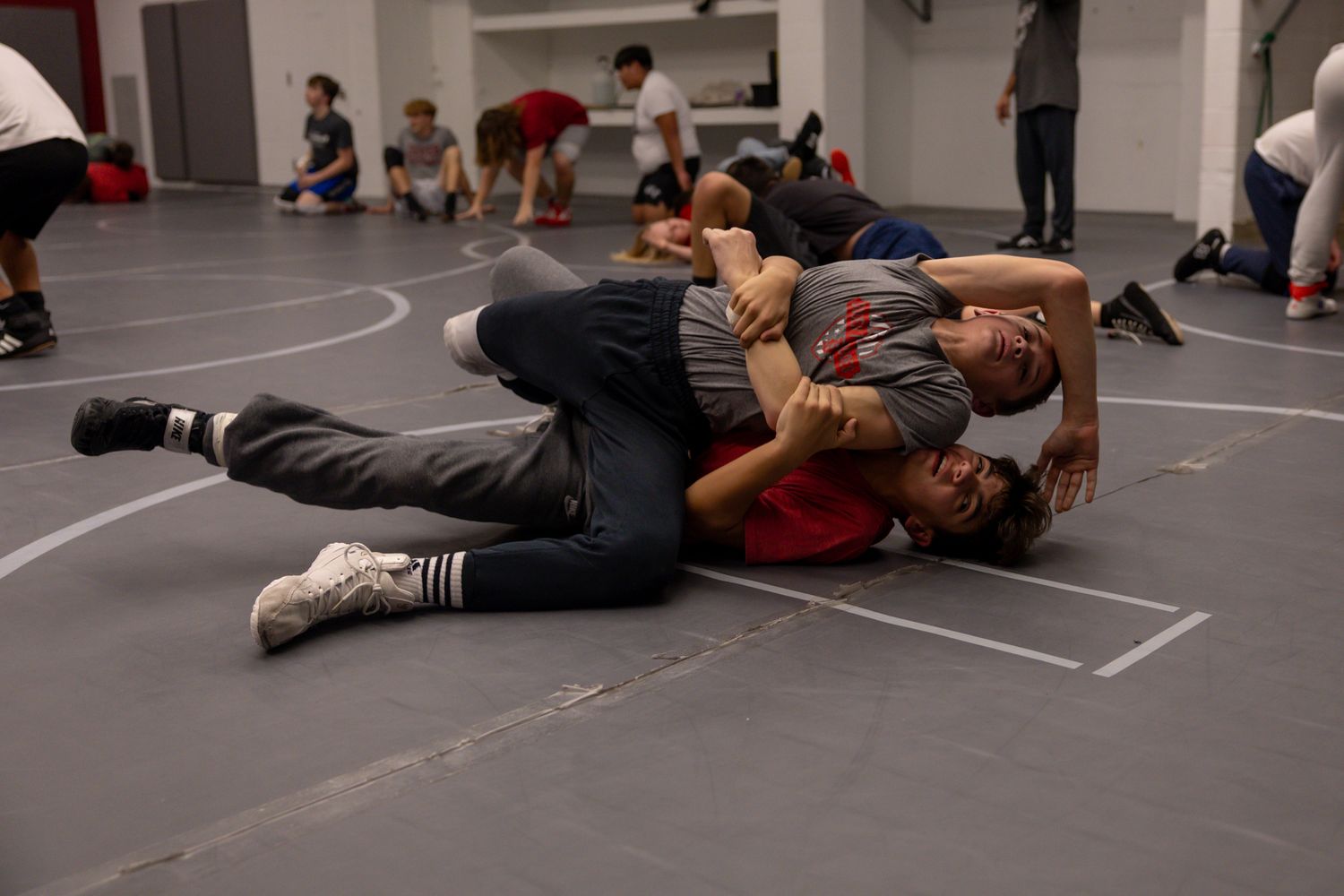 Two wrestlers grapple on a gray mat during a wrestling practice session indoors.