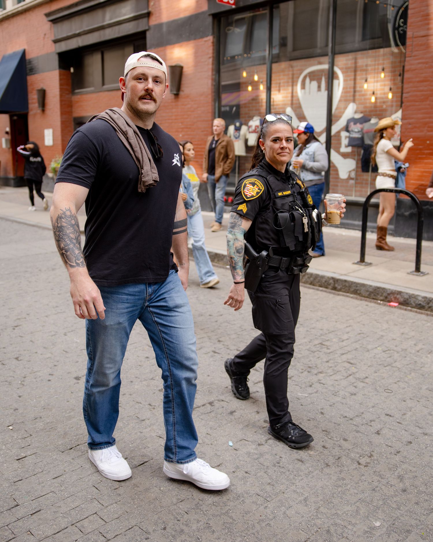 A tall tattooed man in jeans walks beside a female police officer on a brick city street.