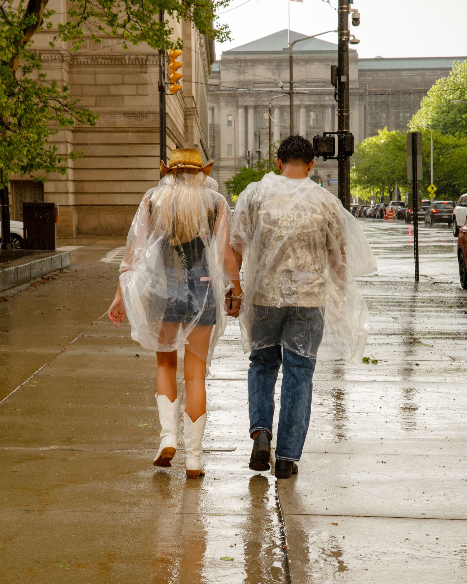 Couple walking hand-in-hand in rain wearing clear plastic ponchos on a wet city sidewalk.