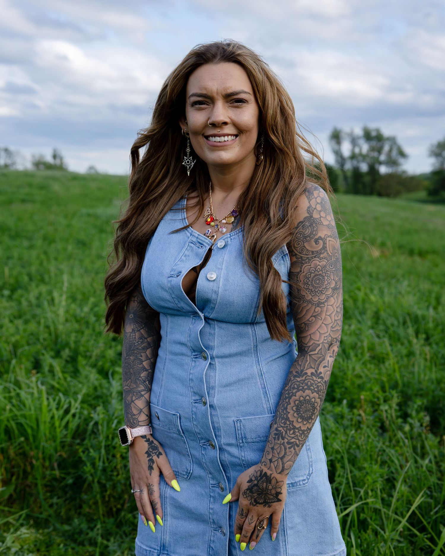 Tattooed woman in denim dress with long wavy hair smiling outdoors in green field, wearing yellow nails and earrings.