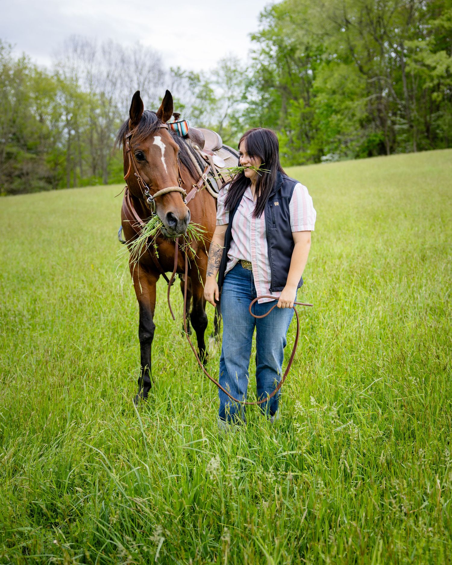A woman in a black vest and jeans walks beside a saddled chestnut horse grazing in a lush green field.