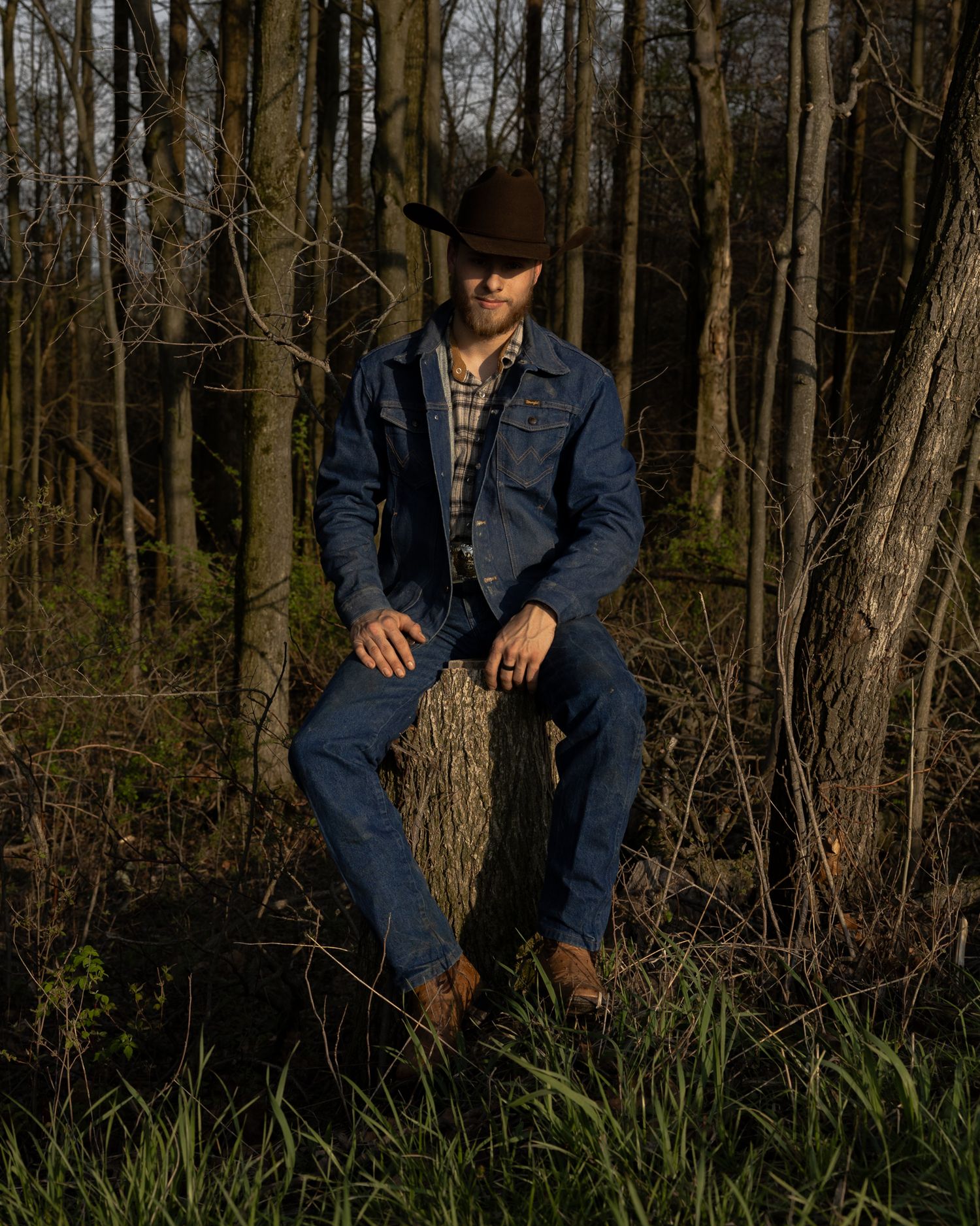 Man in denim jacket and brown cowboy hat poses against trees in golden hour forest light.