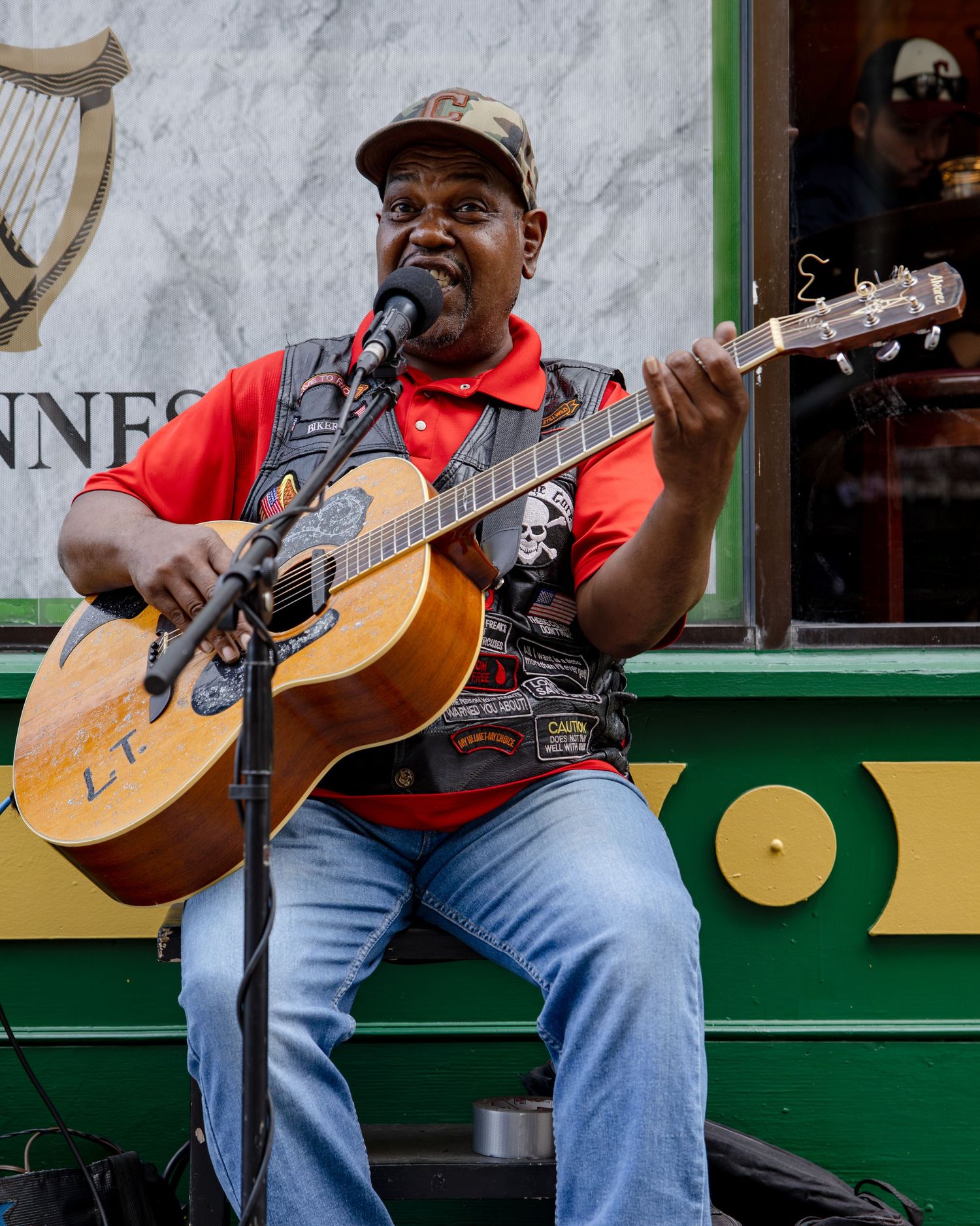A Black musician plays acoustic guitar and sings into a microphone outdoors near a Guinness pub.