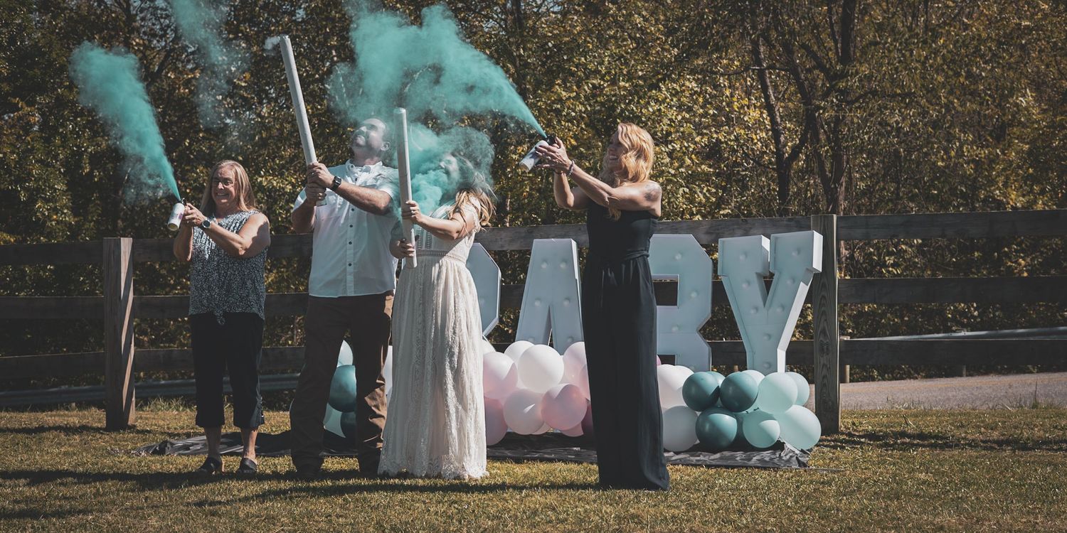Gender reveal party with blue smoke cannons in front of BABY letters and balloons outdoors.