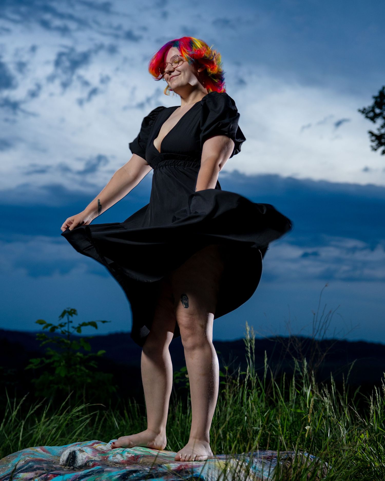 Woman in black dress with colorful hair dancing outdoors at dusk against a dramatic cloudy sky.