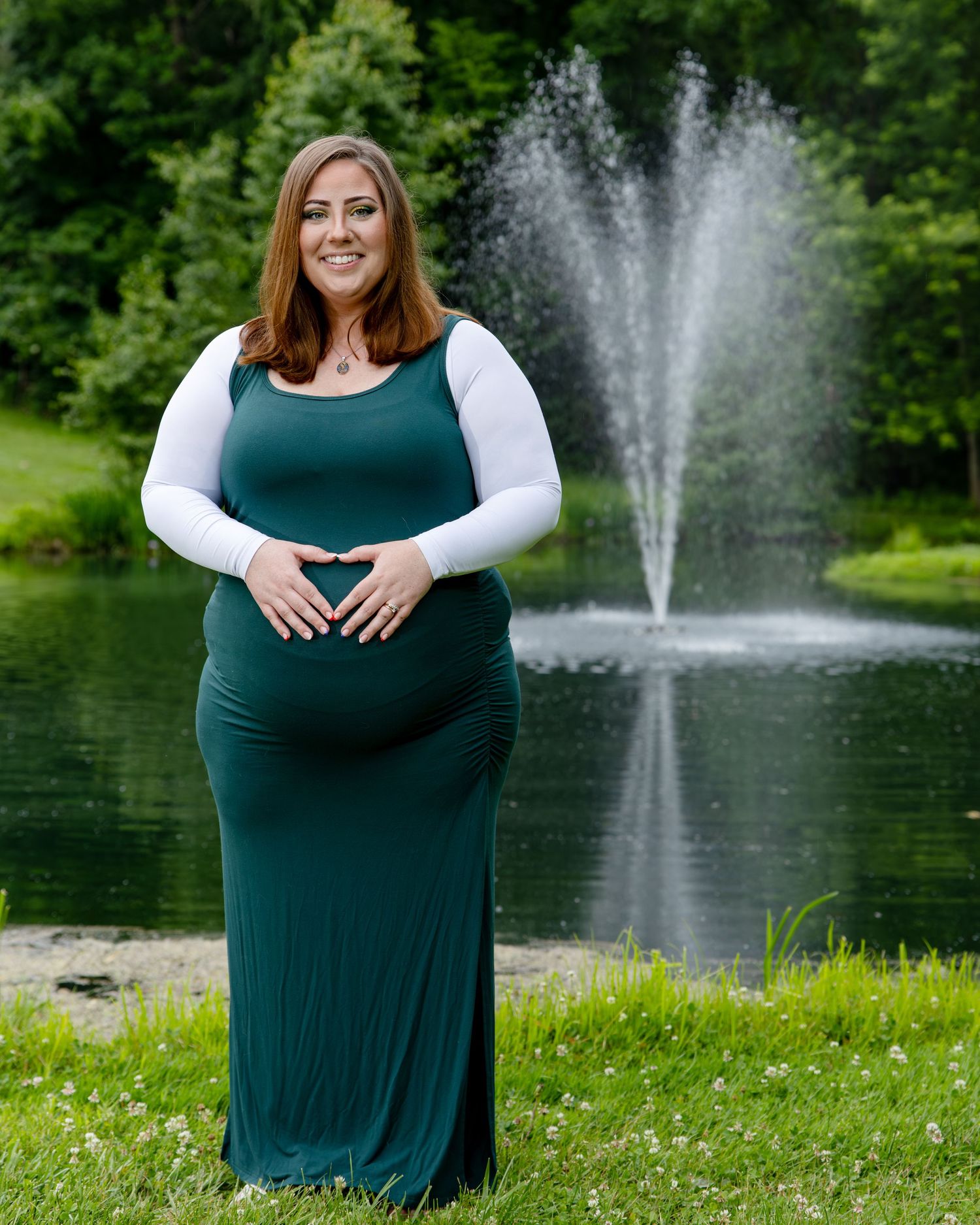 Pregnant woman in teal maxi dress and white top poses by a pond fountain surrounded by lush greenery.