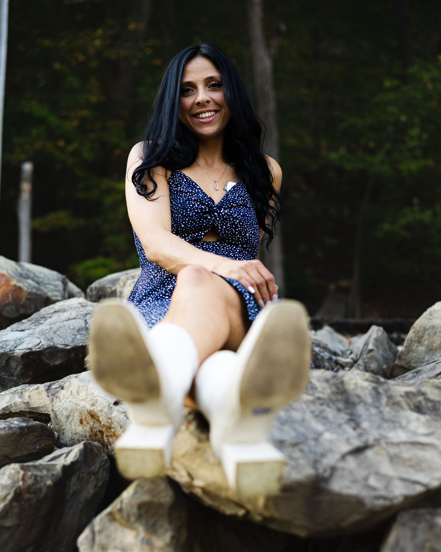 Smiling brunette woman in blue floral dress sitting on rocks with white boots.