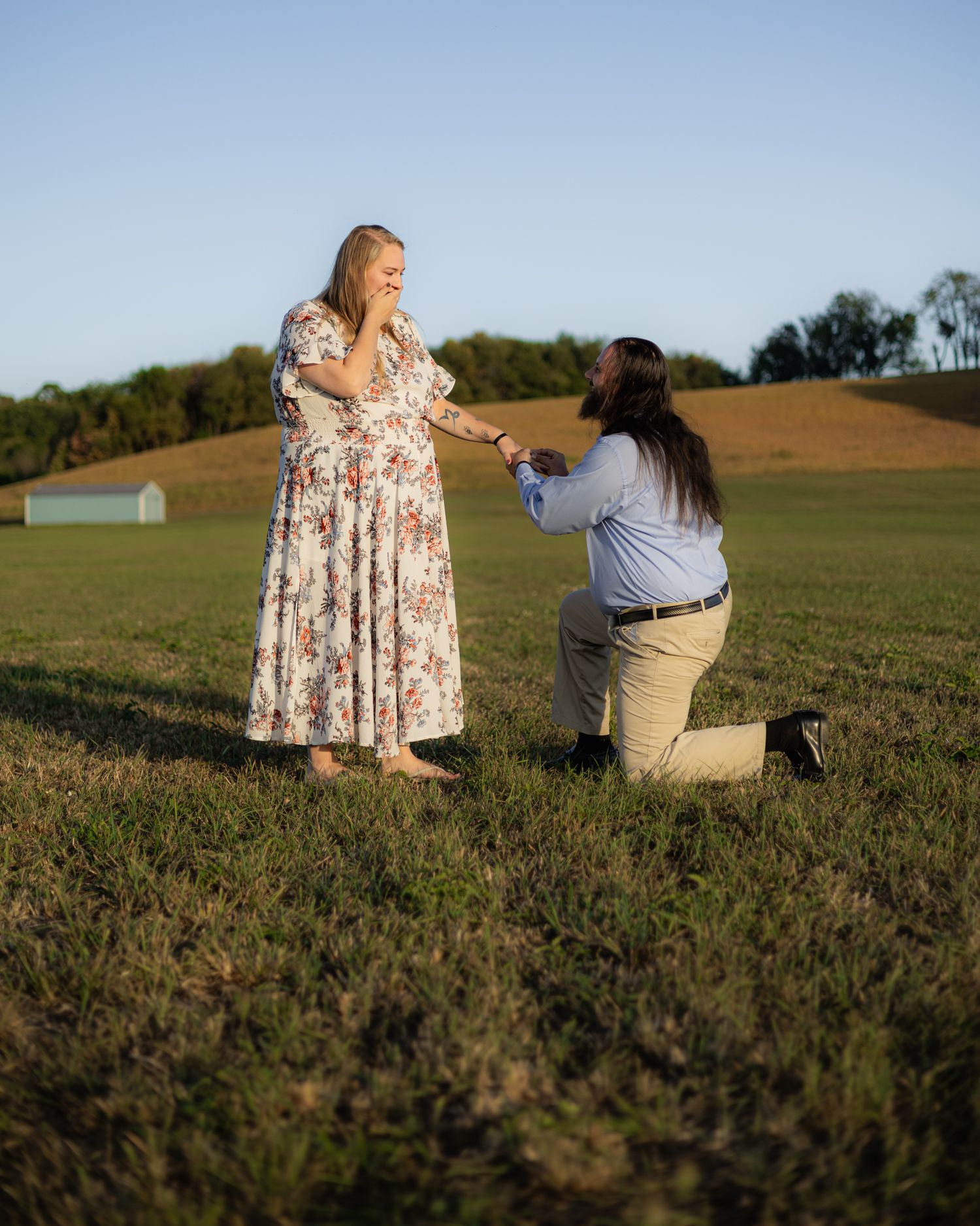 Person kneeling on one knee proposing to woman in floral dress in open field at sunset.