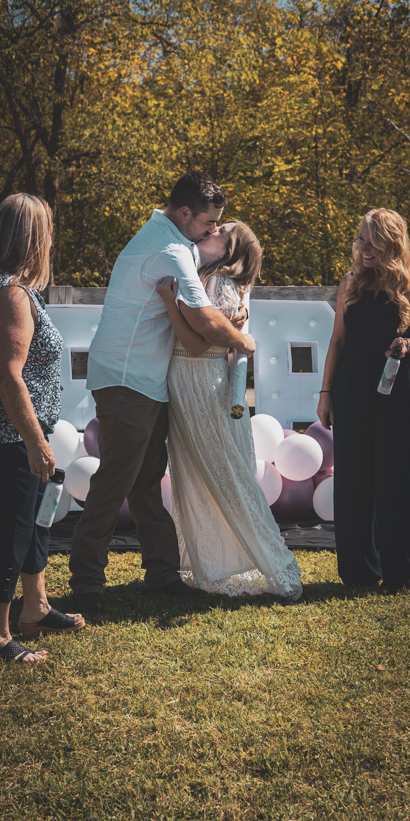 Couple shares a romantic kiss at an outdoor gender reveal party with pink and white balloons in the background.