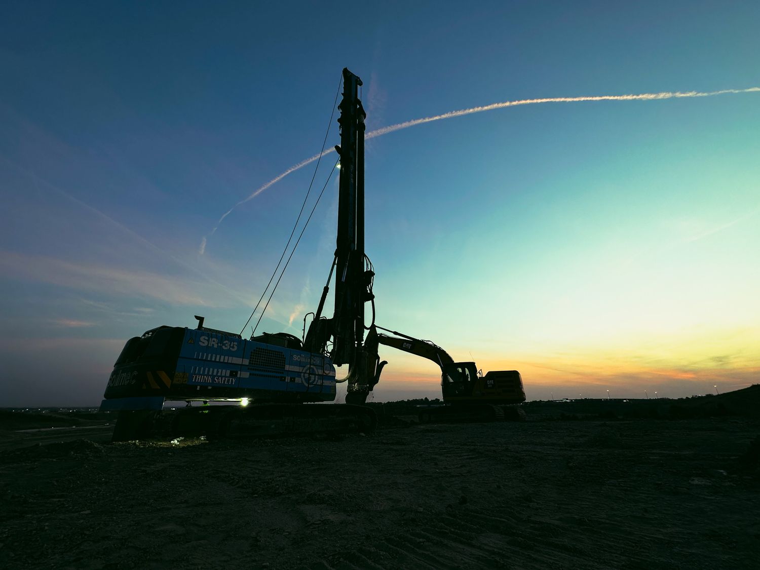 Drilling rig silhouette against a vibrant sunset sky with contrail streaks at a construction site.