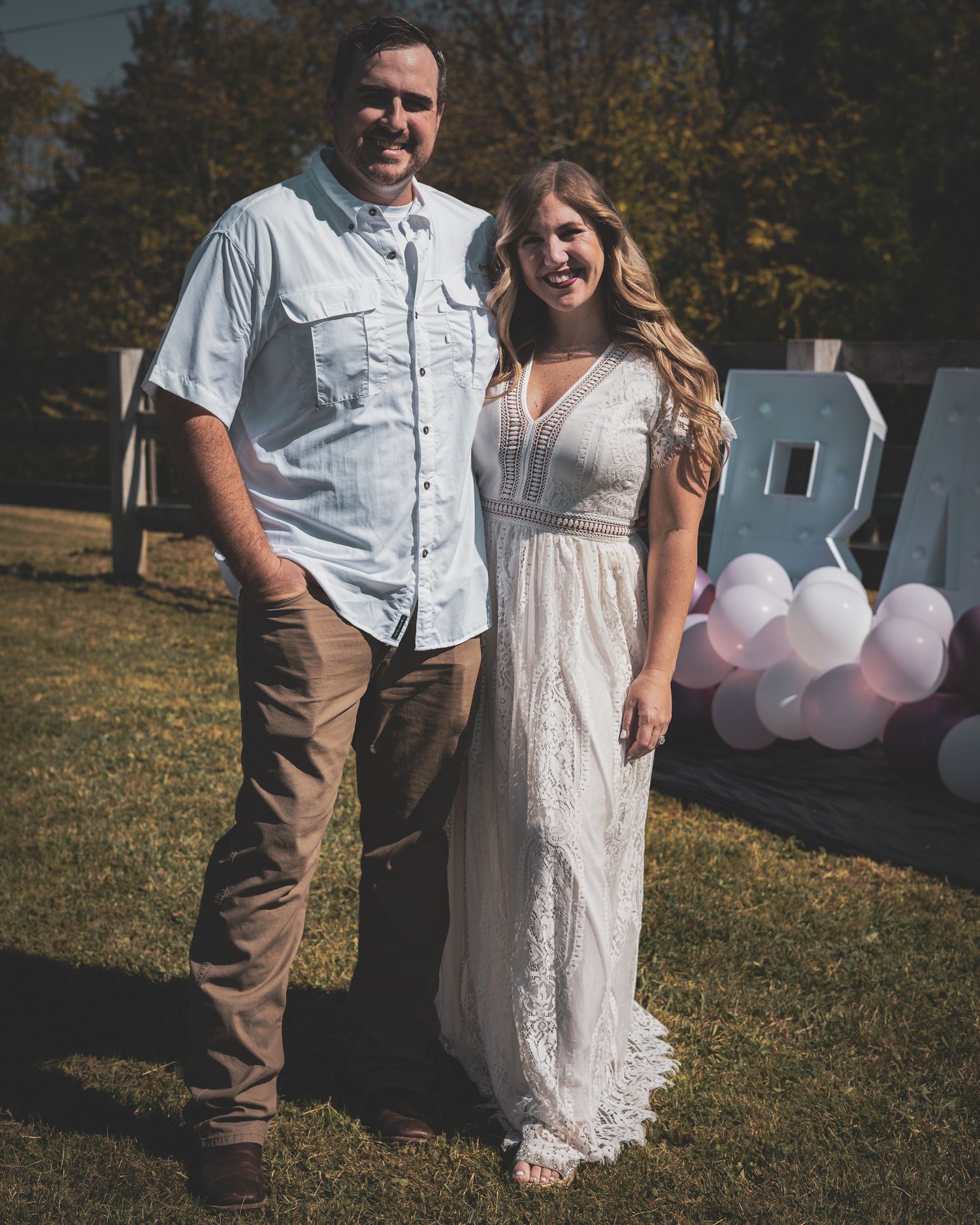 Expecting couple posing in front of BABY balloon display at outdoor gender reveal.