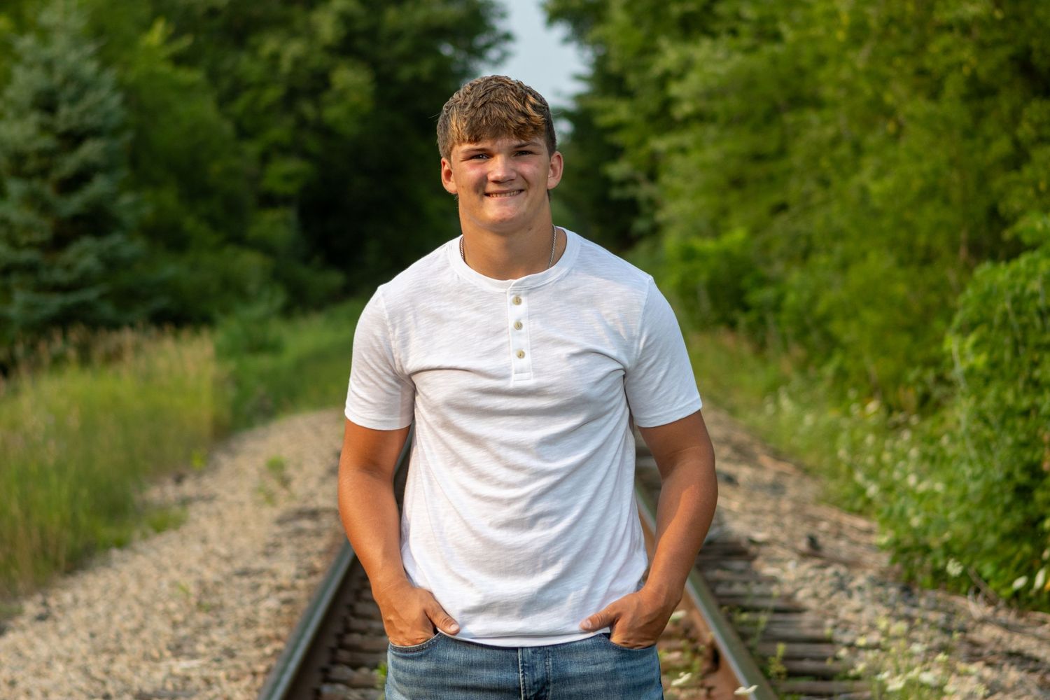 Young man in white t-shirt and jeans standing on railroad tracks surrounded by lush green trees.