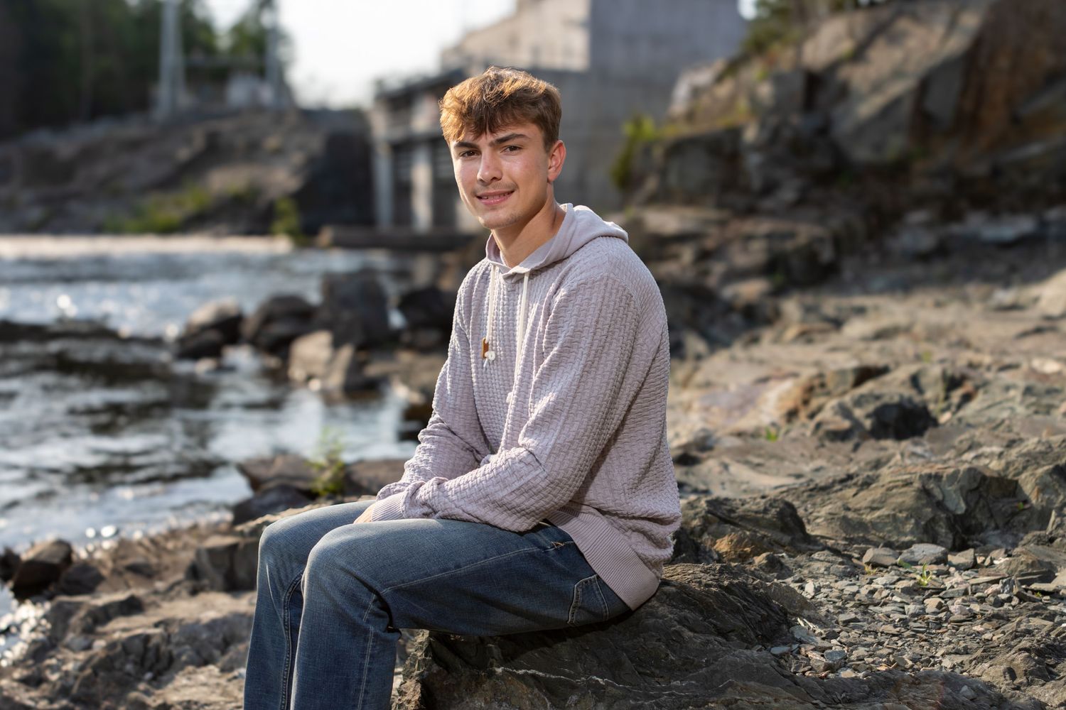 A young man in a beige hoodie and jeans sits on rocky shoreline with water and stone structures in background.