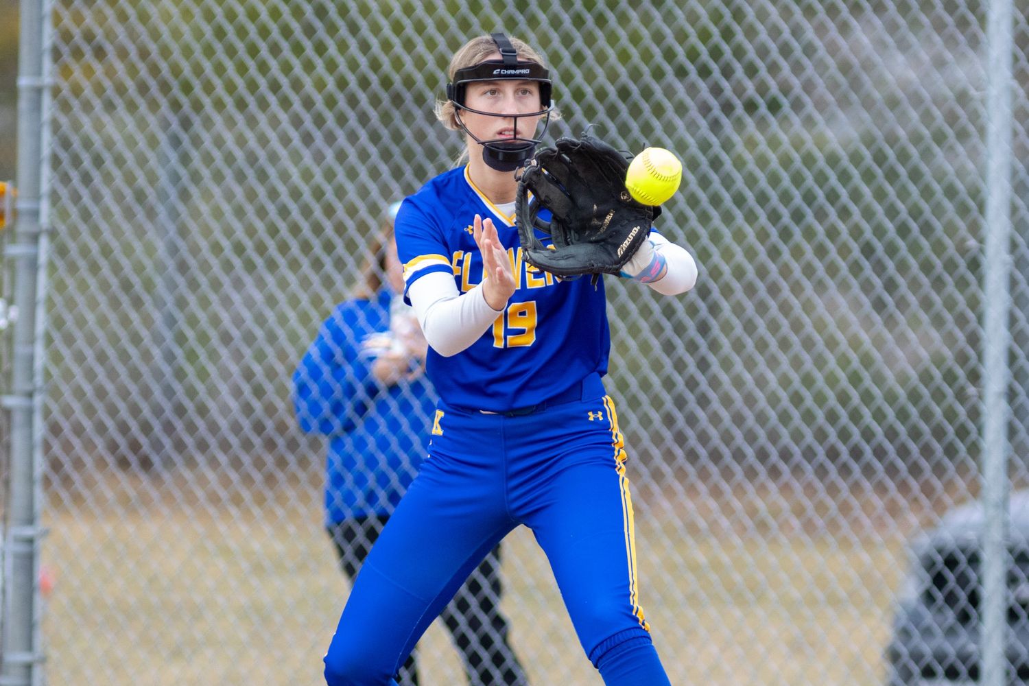 Softball player in blue and gold uniform catching a yellow ball with glove during an outdoor game.