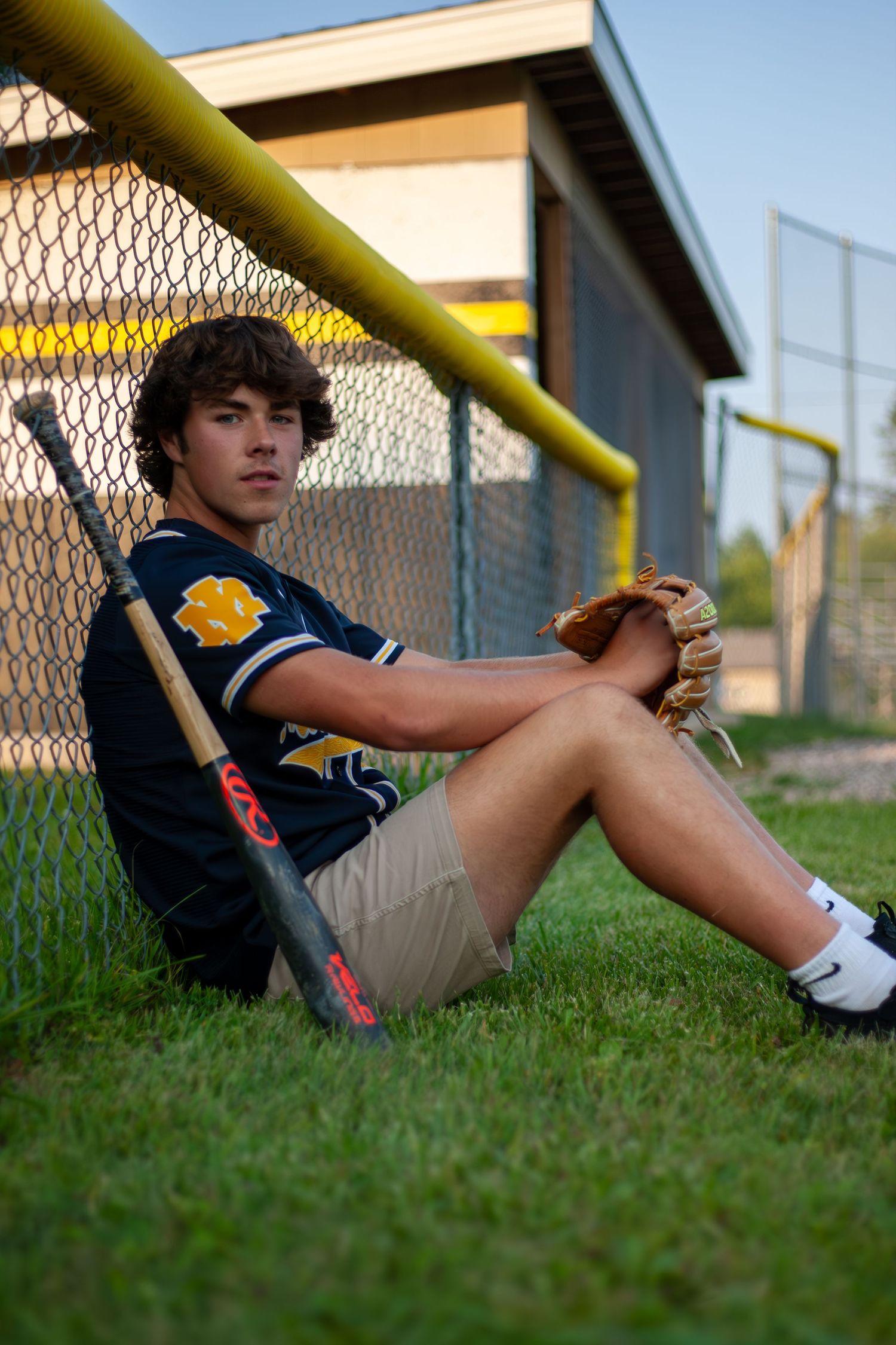 Baseball player rests against chain link fence with bat and glove during practice.