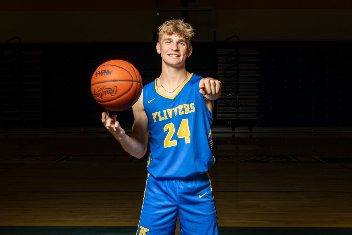 Young male basketball player in blue Flyers #24 jersey poses with ball on gym court.