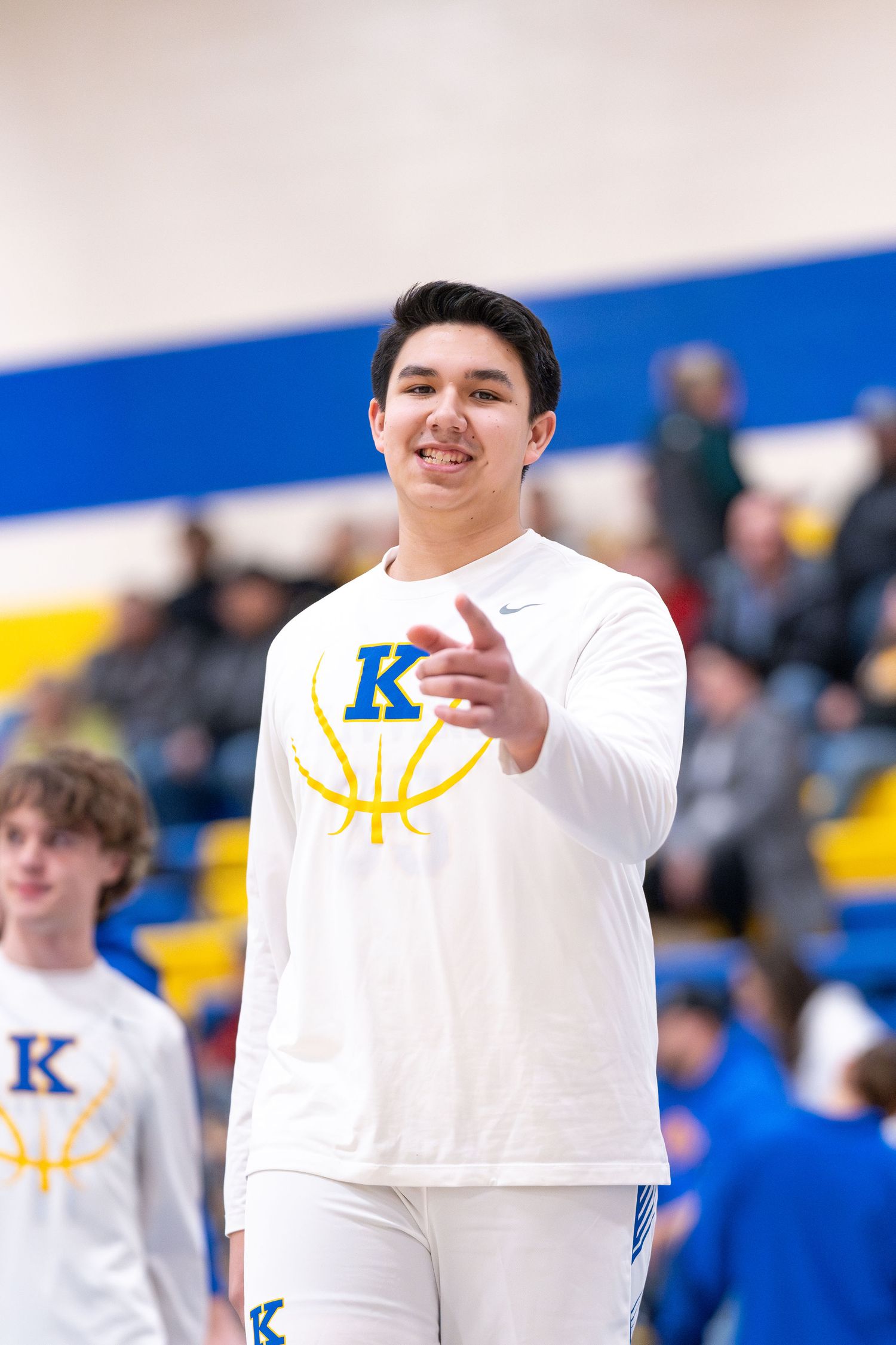 Basketball fan in white shirt with K logo gestures in gymnasium bleachers.