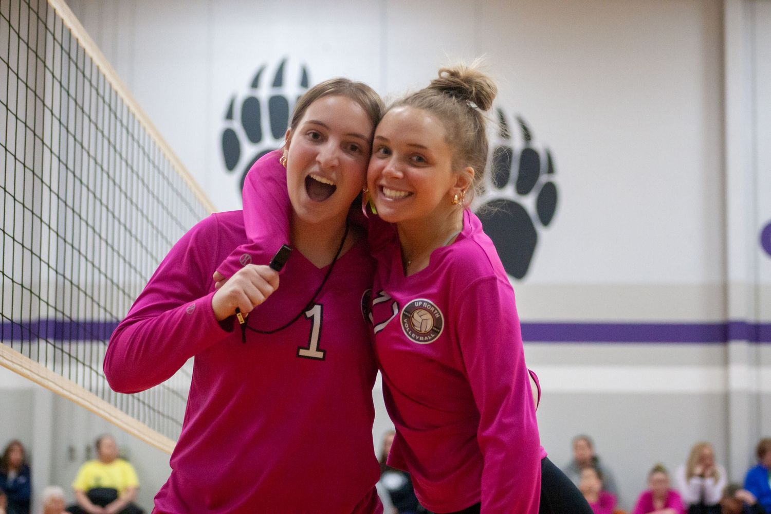 Two volleyball players in pink jerseys celebrate during match near paw print logo.