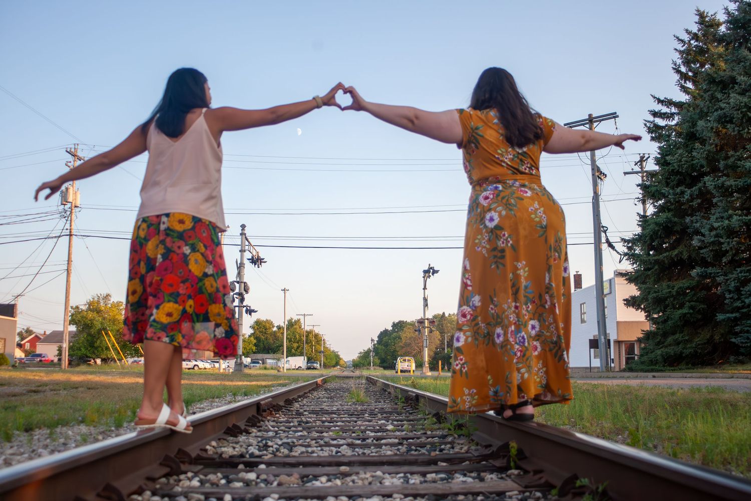 Friends in yellow dresses making heart shapes with arms on train tracks at sunset.