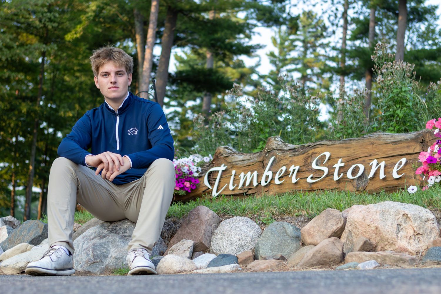 Young man in blue hoodie sitting on rocks beside a Timber Stone wooden sign surrounded by flowers and trees.