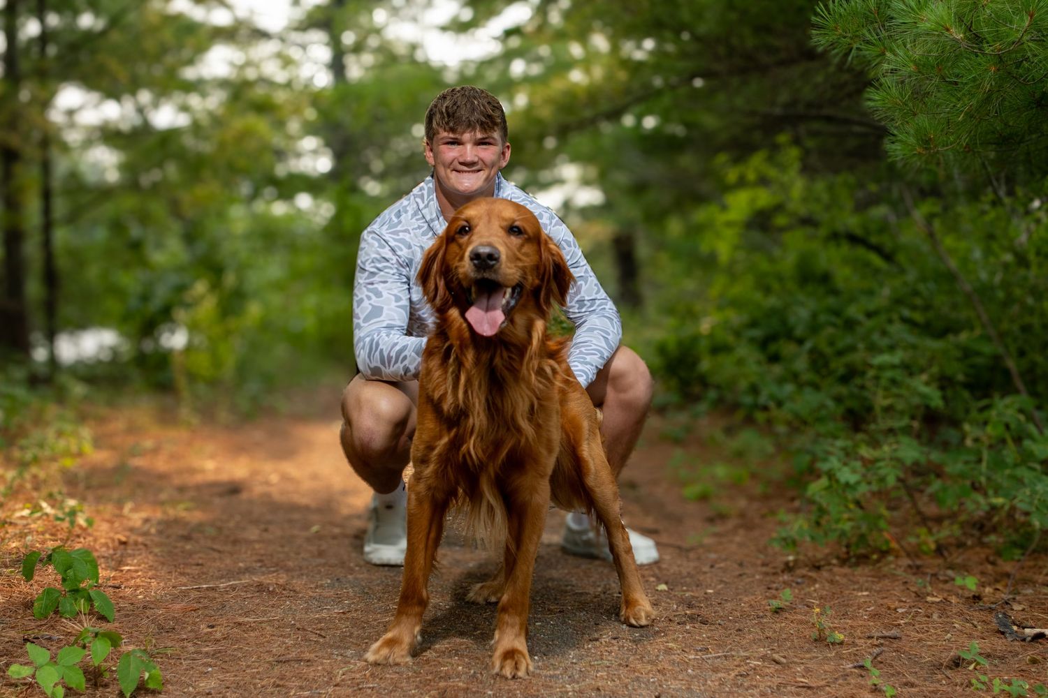 A smiling person crouches beside a golden Irish Setter dog on a forest trail surrounded by lush green trees.