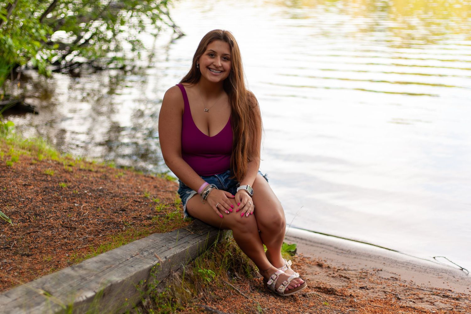 Person in burgundy top sits on stone ledge near tranquil lake at sunset.