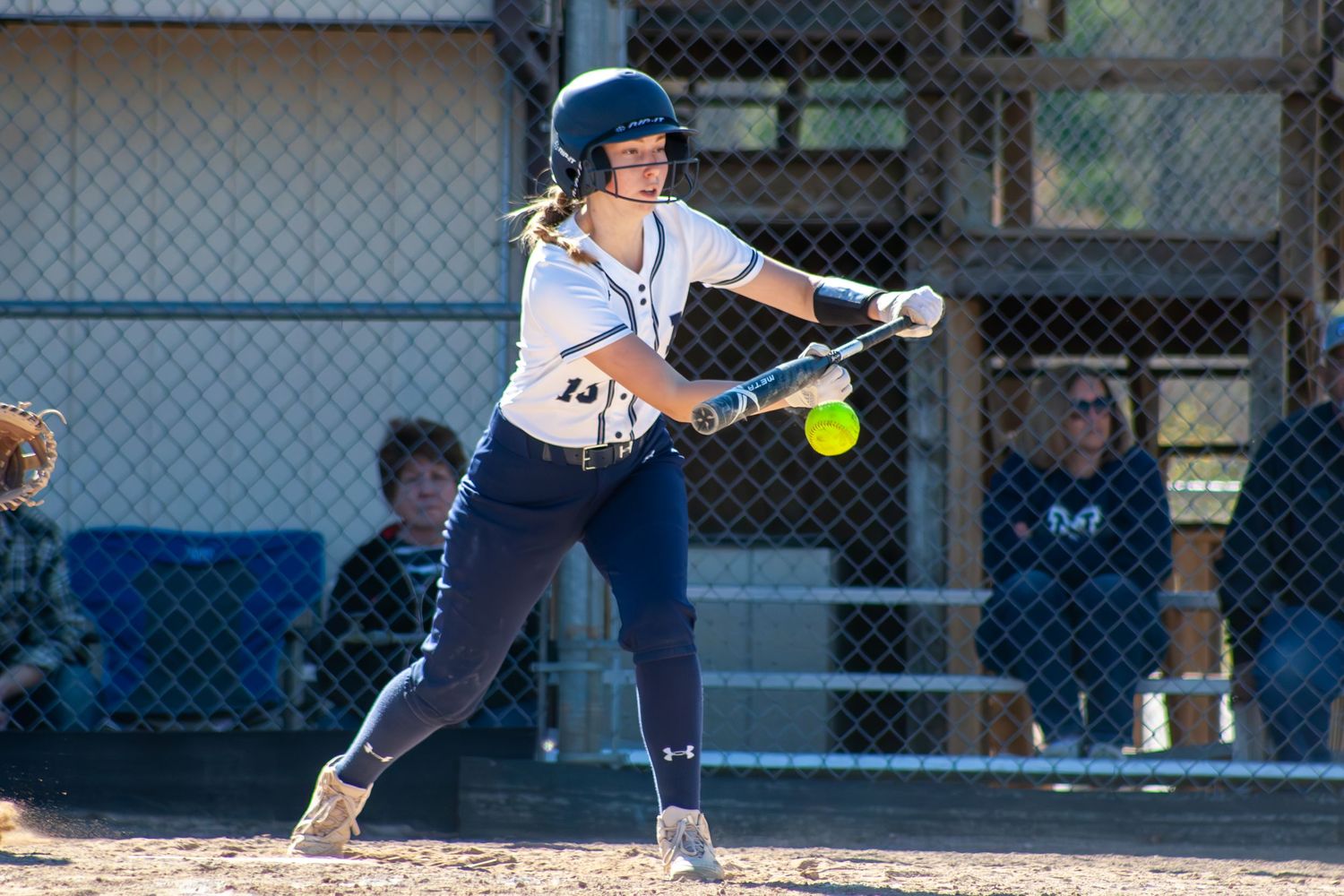 Softball batter in navy uniform swings at incoming pitch during game.