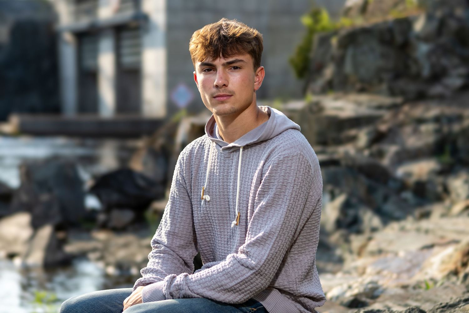 A young man in a gray hoodie sits on rocks outdoors, with a blurred stone structure in the background.