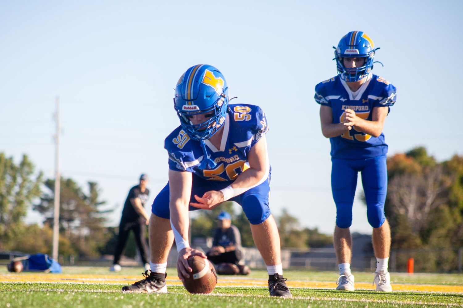 Football players in blue uniforms practicing field goal kicks on a sunny day.