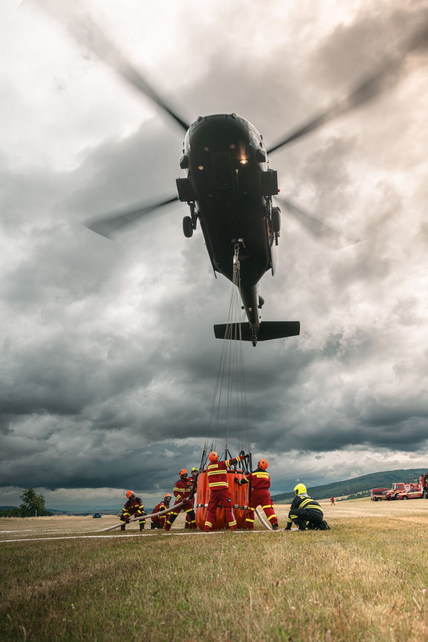 Military helicopter hovering over field equipment under dramatic stormy skies.