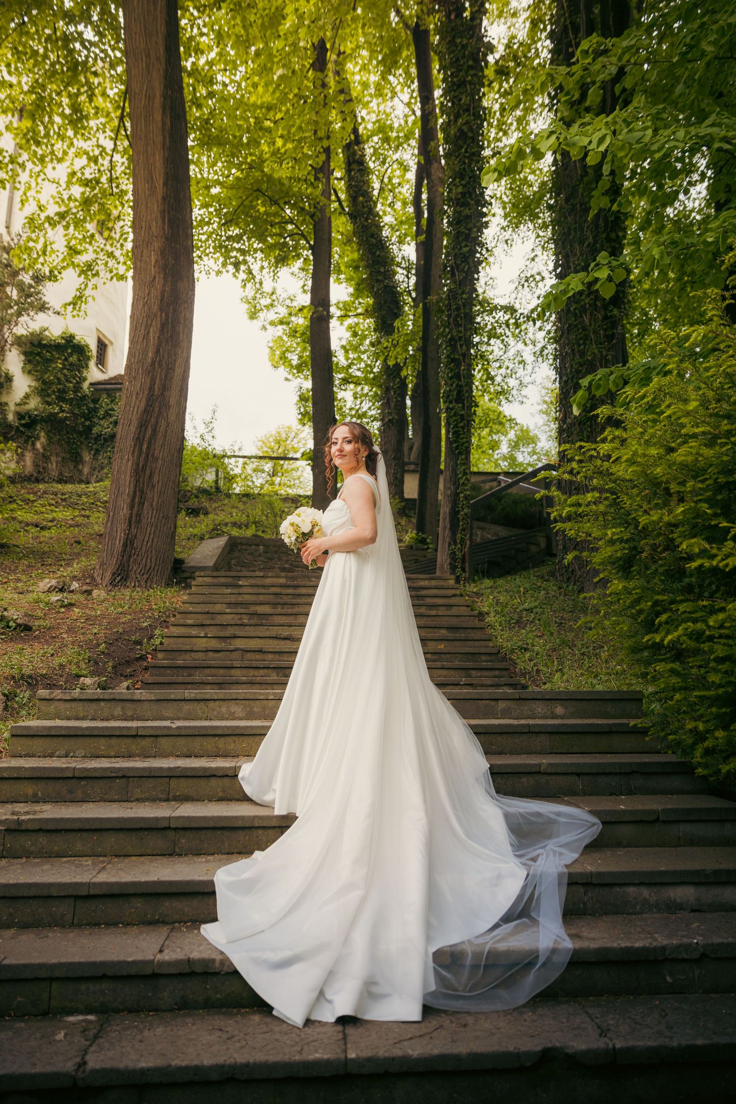 White wedding dress flowing on stone steps surrounded by lush green trees.
