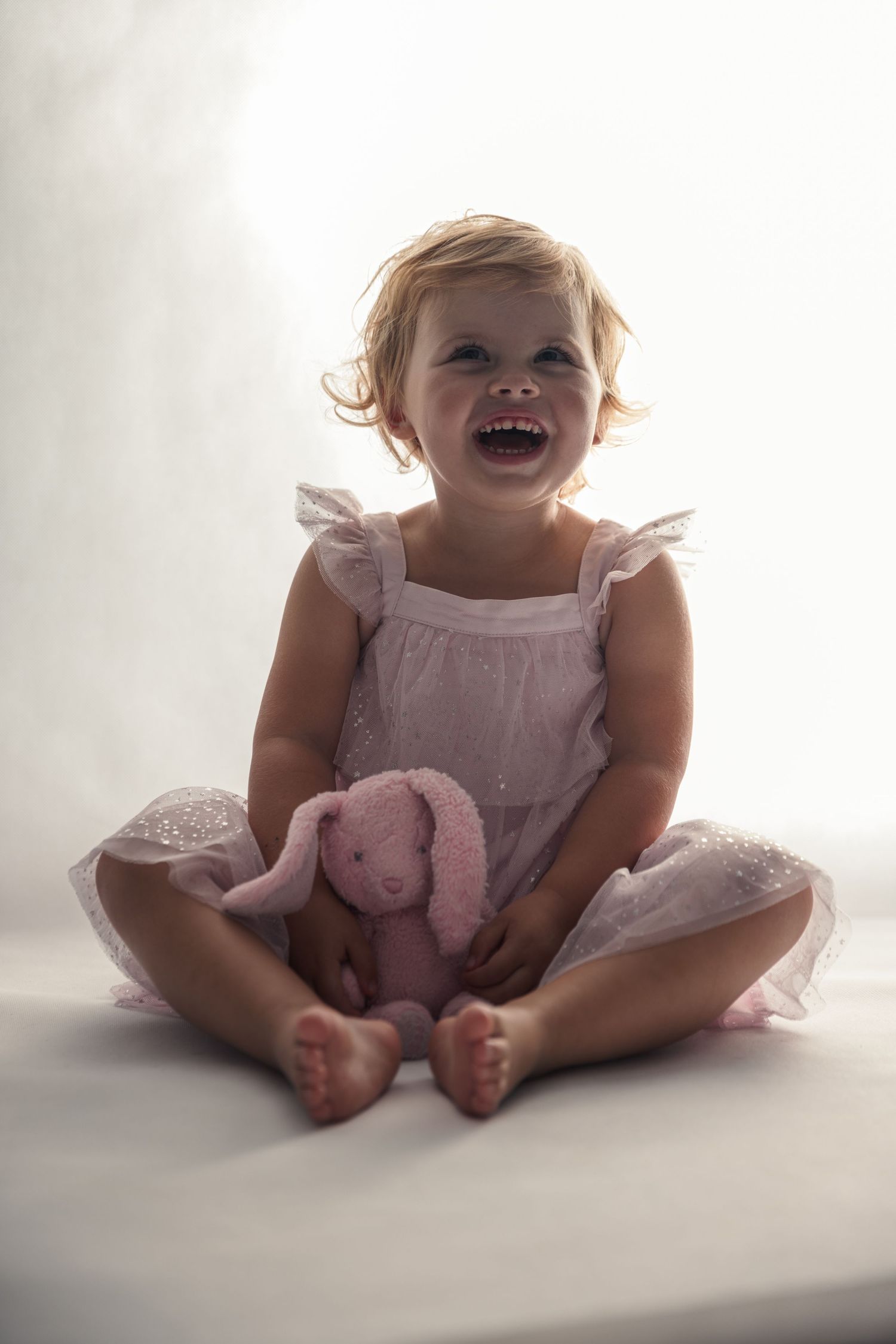 Joyful toddler in pink dress sits with stuffed bunny toy on white background.