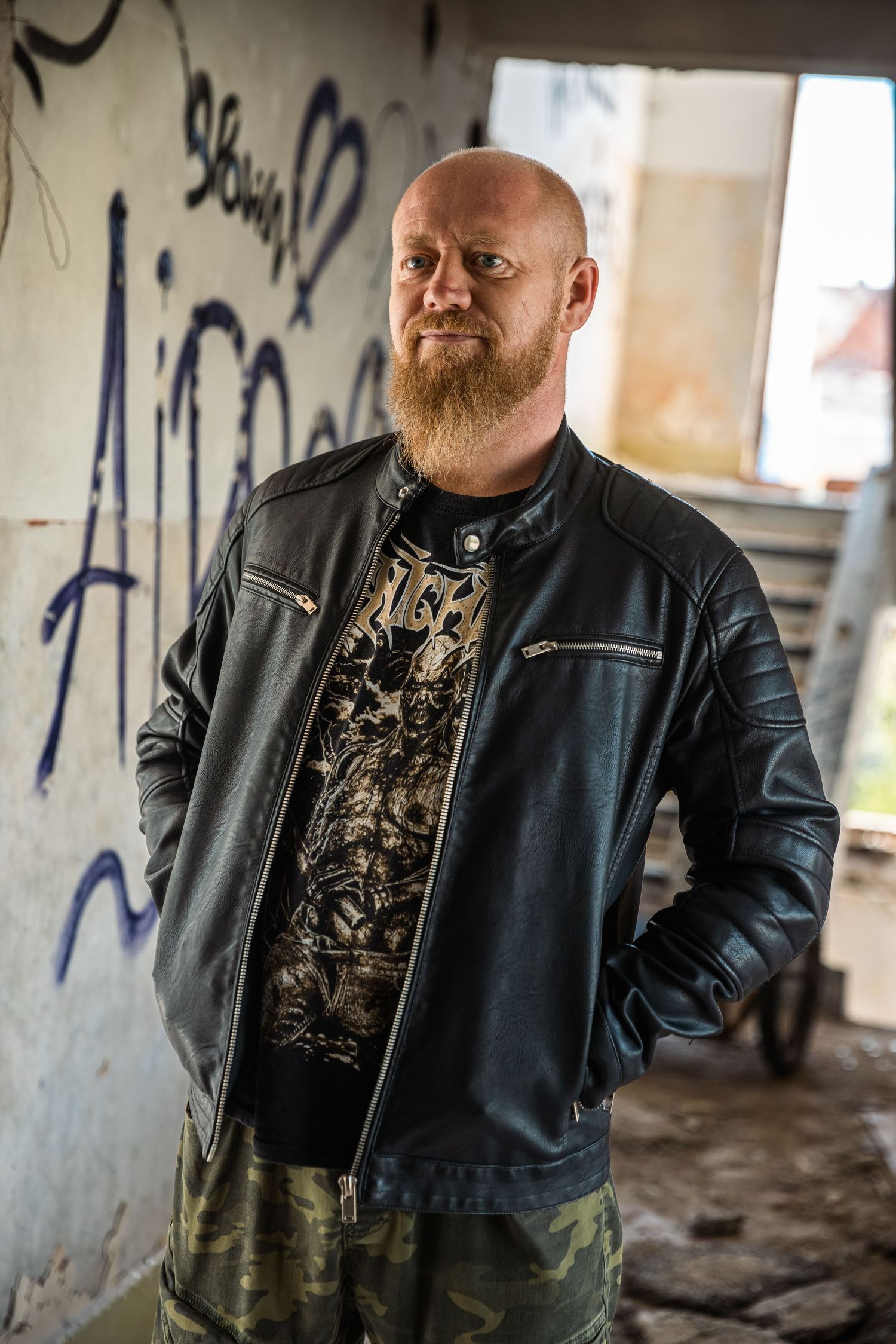 Person wearing leather jacket and dark clothing stands against graffiti wall in abandoned building.