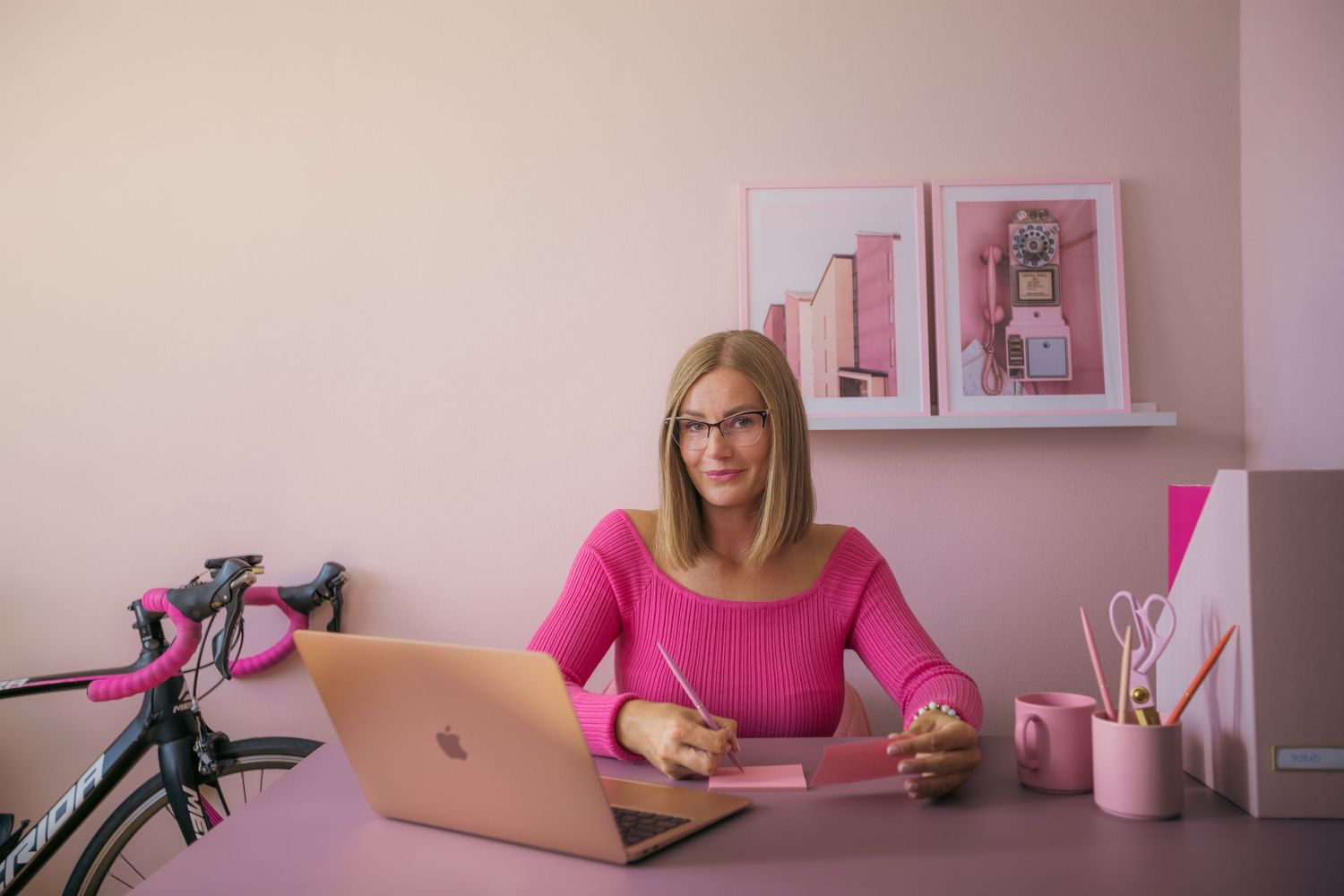 Woman in pink top works at desk with laptop in feminine pink office space.