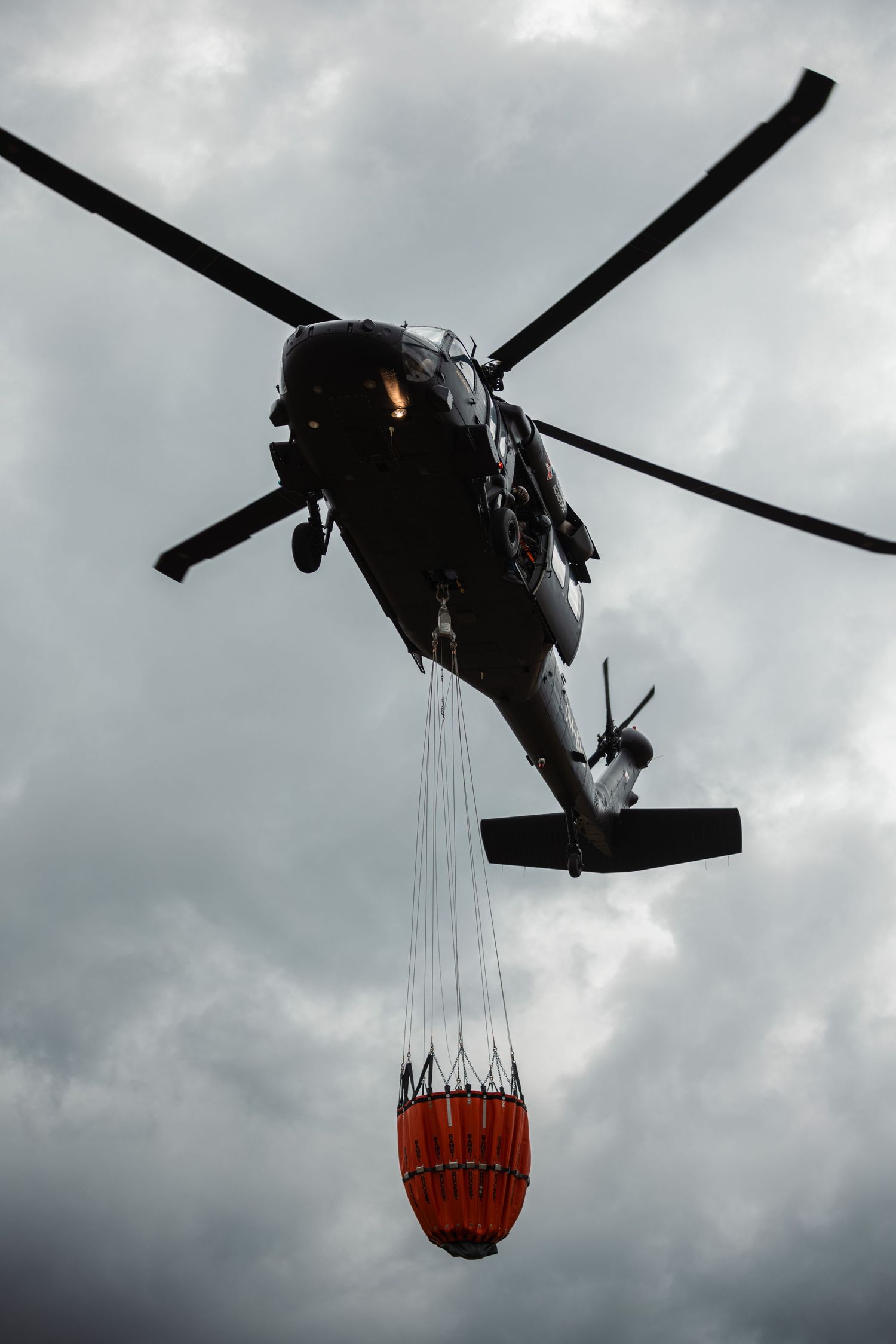 Military helicopter carrying a large orange water bucket during firefighting operations against cloudy skies.