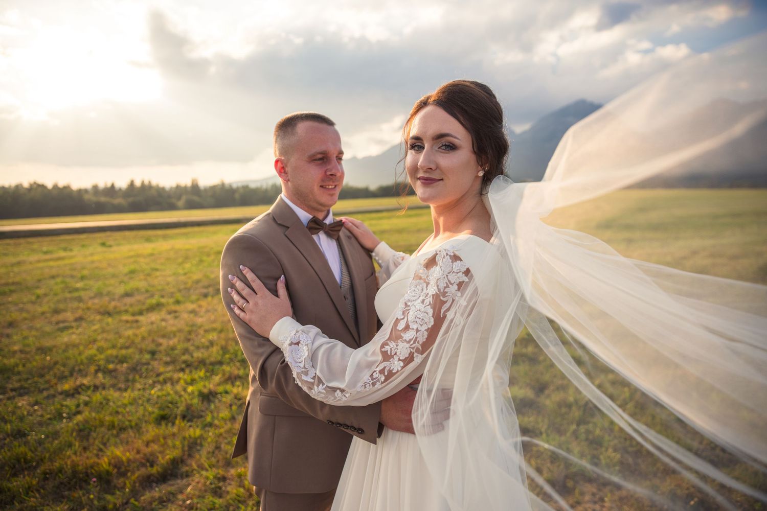 Wedding couple embraces in scenic meadow with flowing white veil and lace dress details.