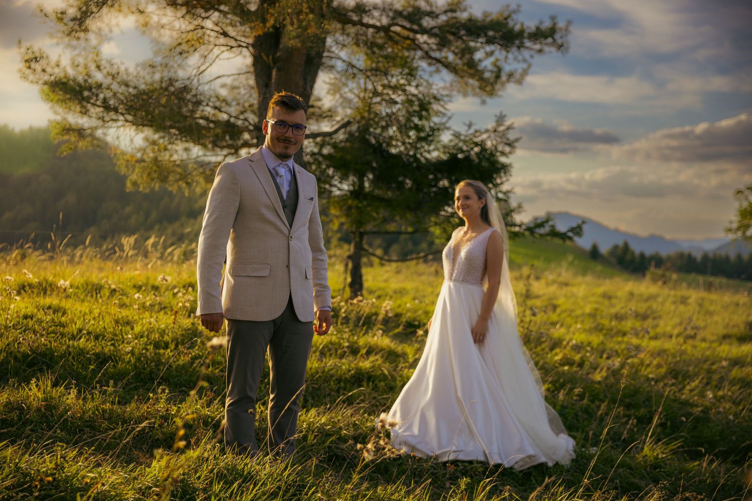 A couple walks through a sunlit meadow with mountains in the distance.