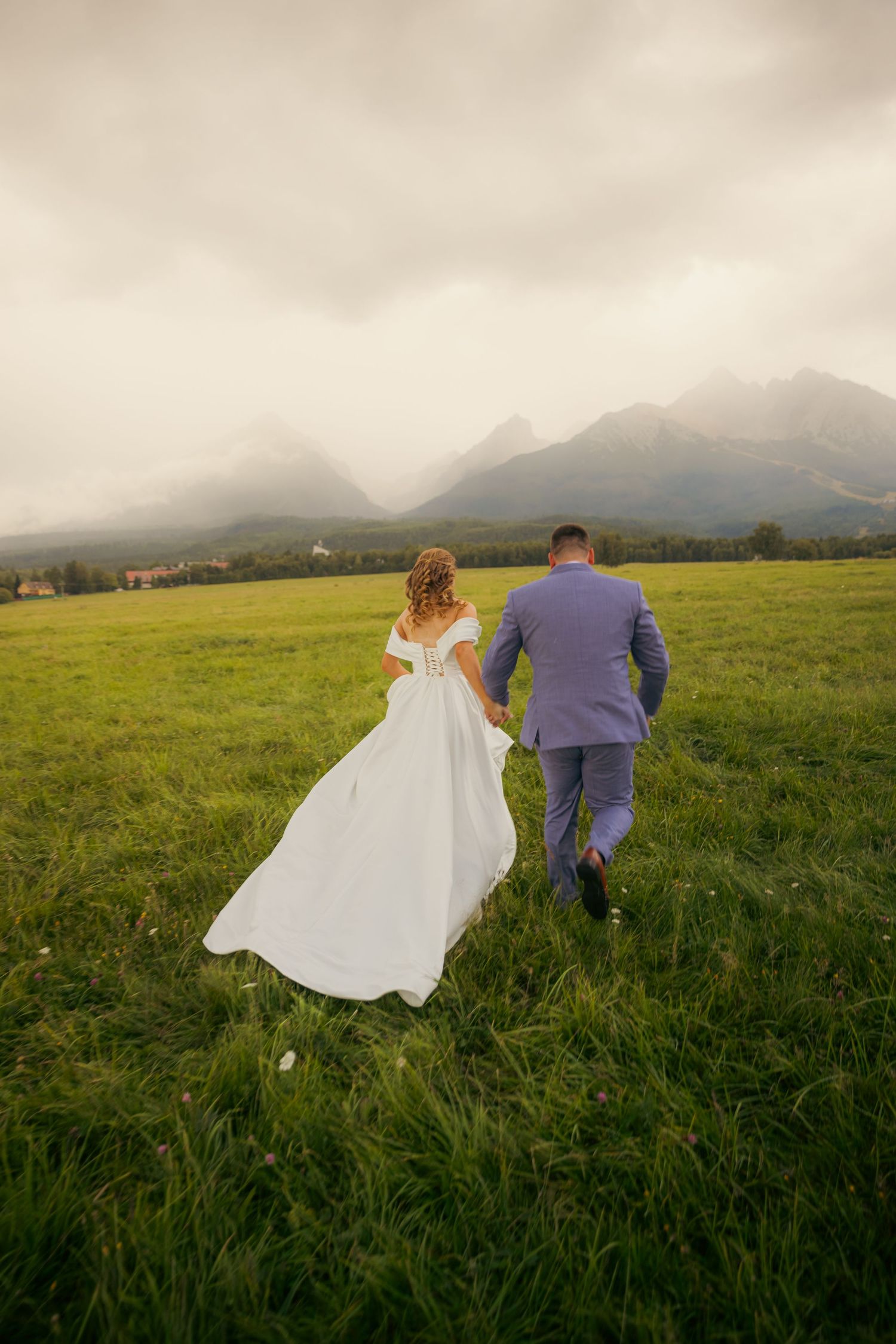 Bride and groom walking through grassy field with misty mountains behind them.