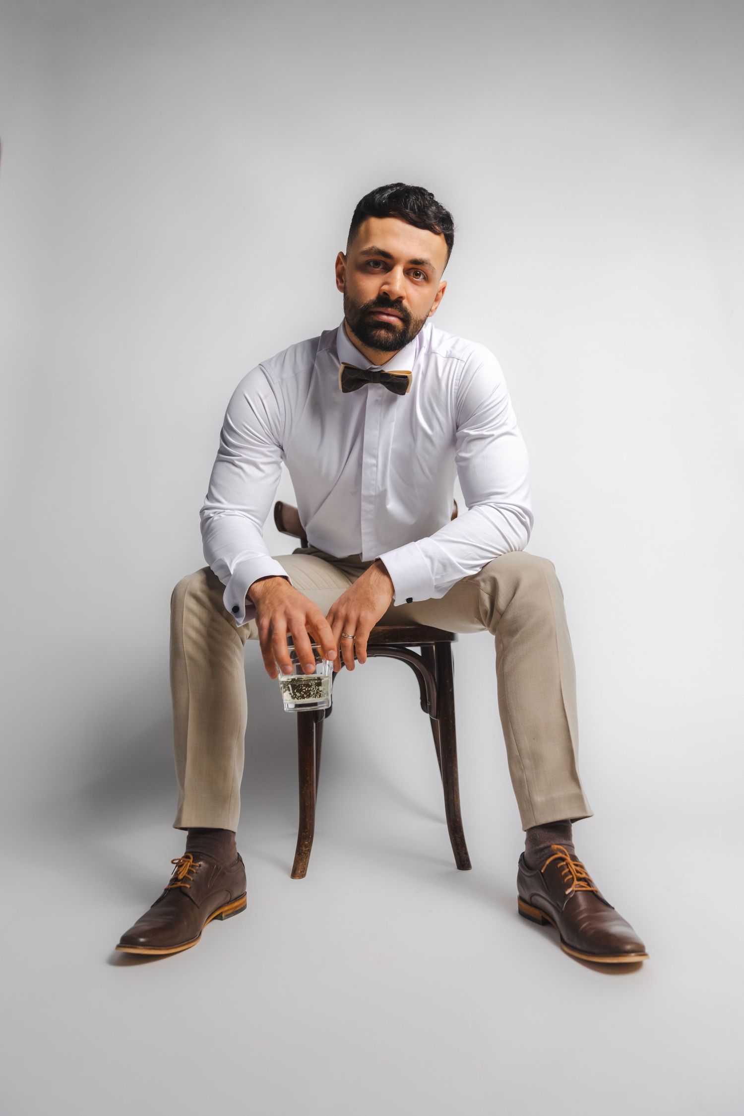 Stylish groom in beige suit and bowtie poses on wooden chair against white backdrop.