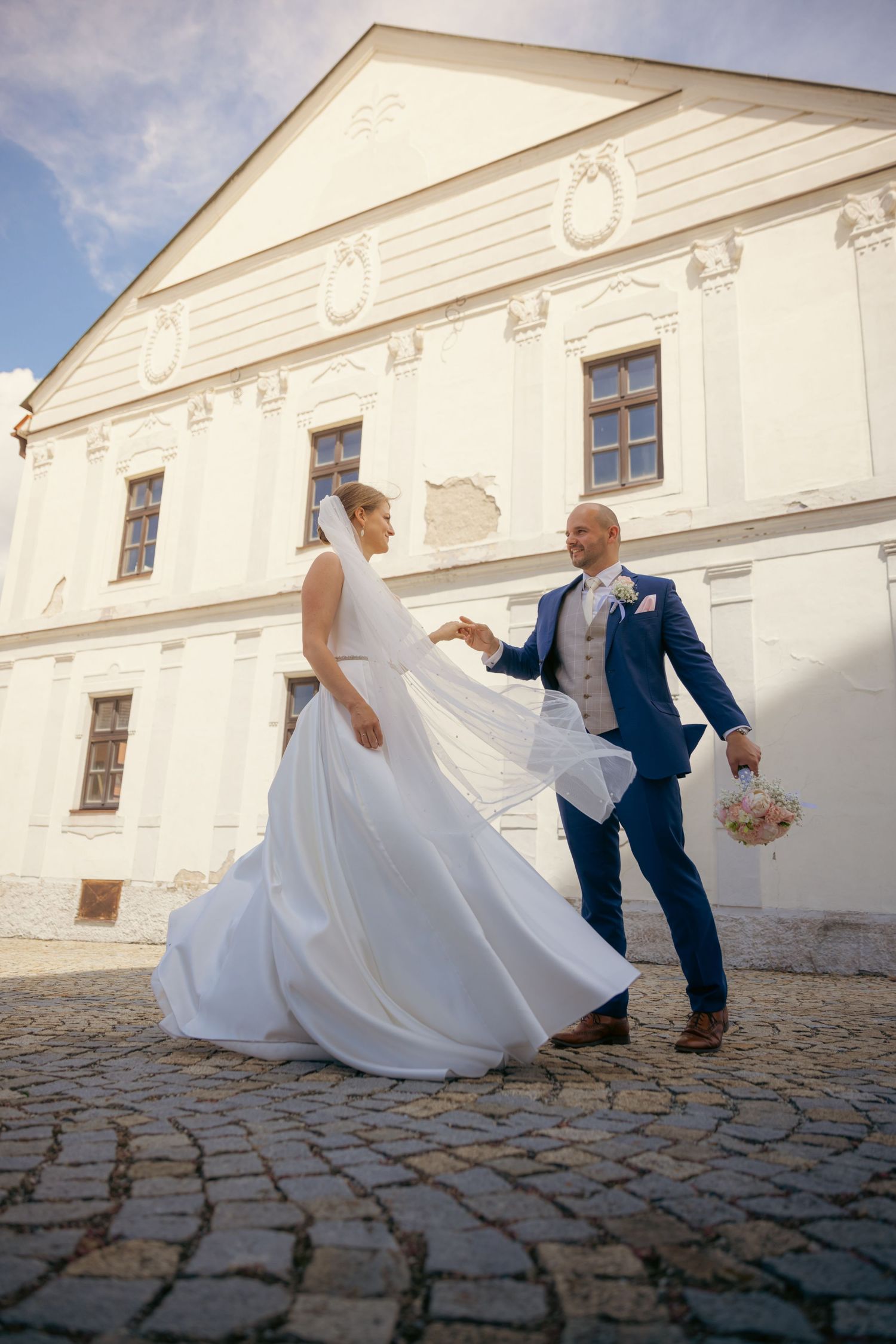 Wedding couple in elegant attire dancing playfully against a white historic building backdrop.