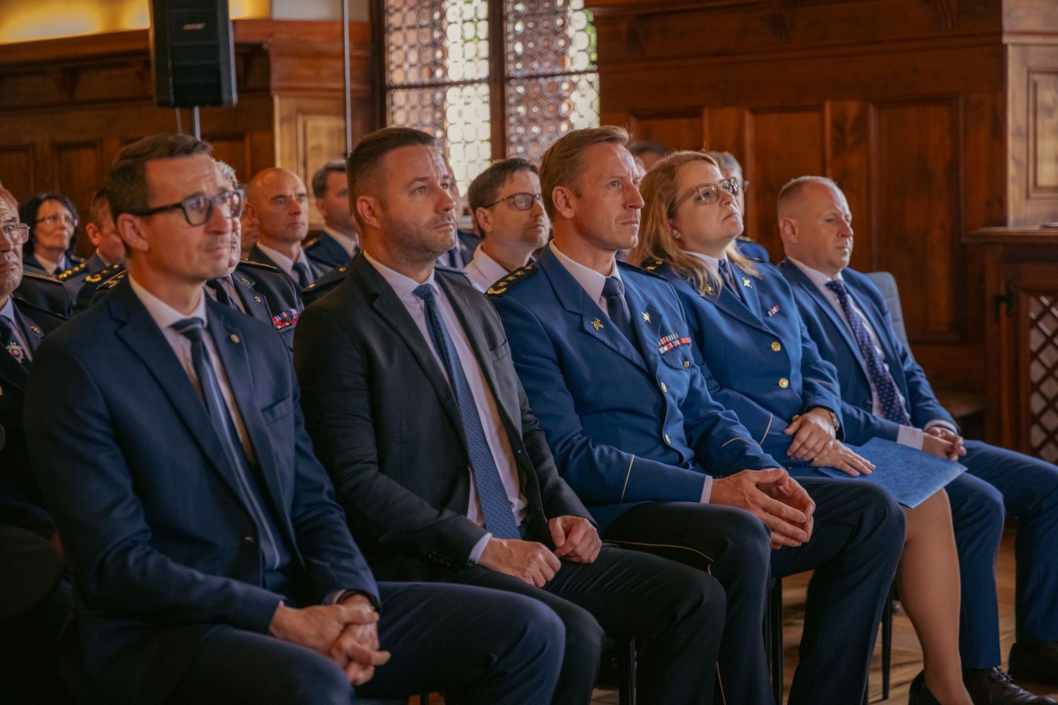 Formal gathering in a wood-paneled room with attendees in business attire seated in rows.