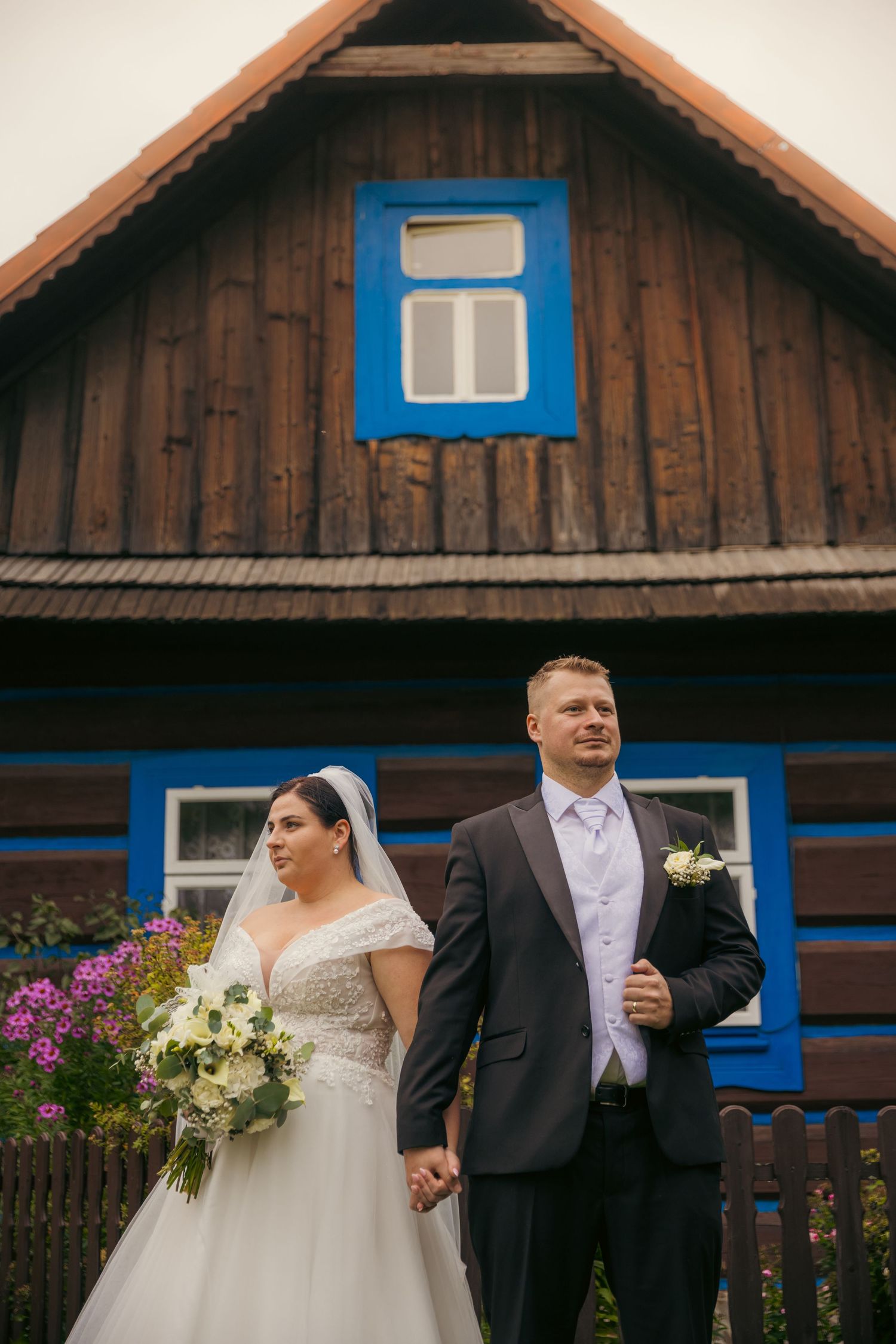 A wedding couple poses in front of a rustic wooden cabin with blue trim and flowers.