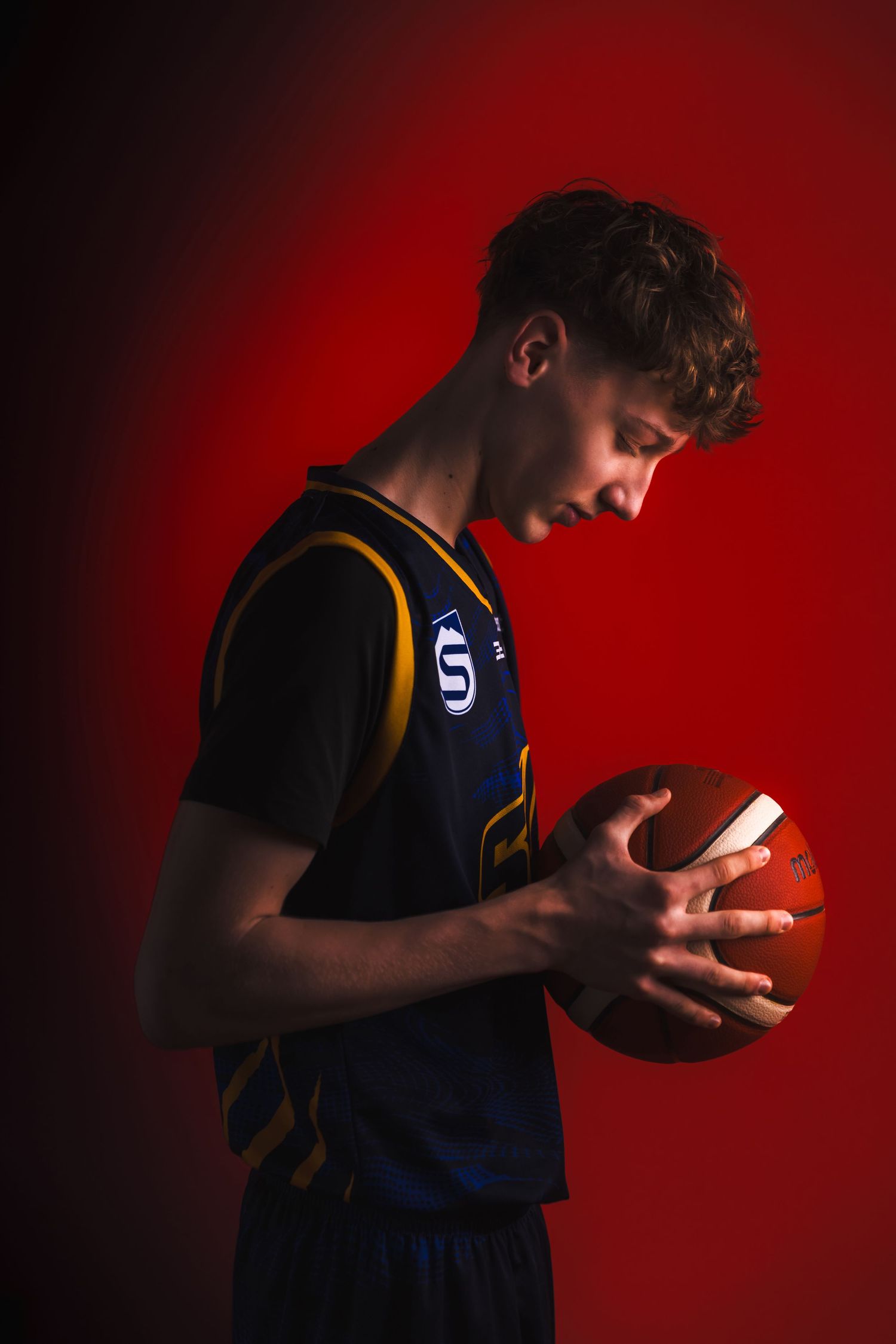 Basketball player examines ball against red backdrop in moody sports portrait.