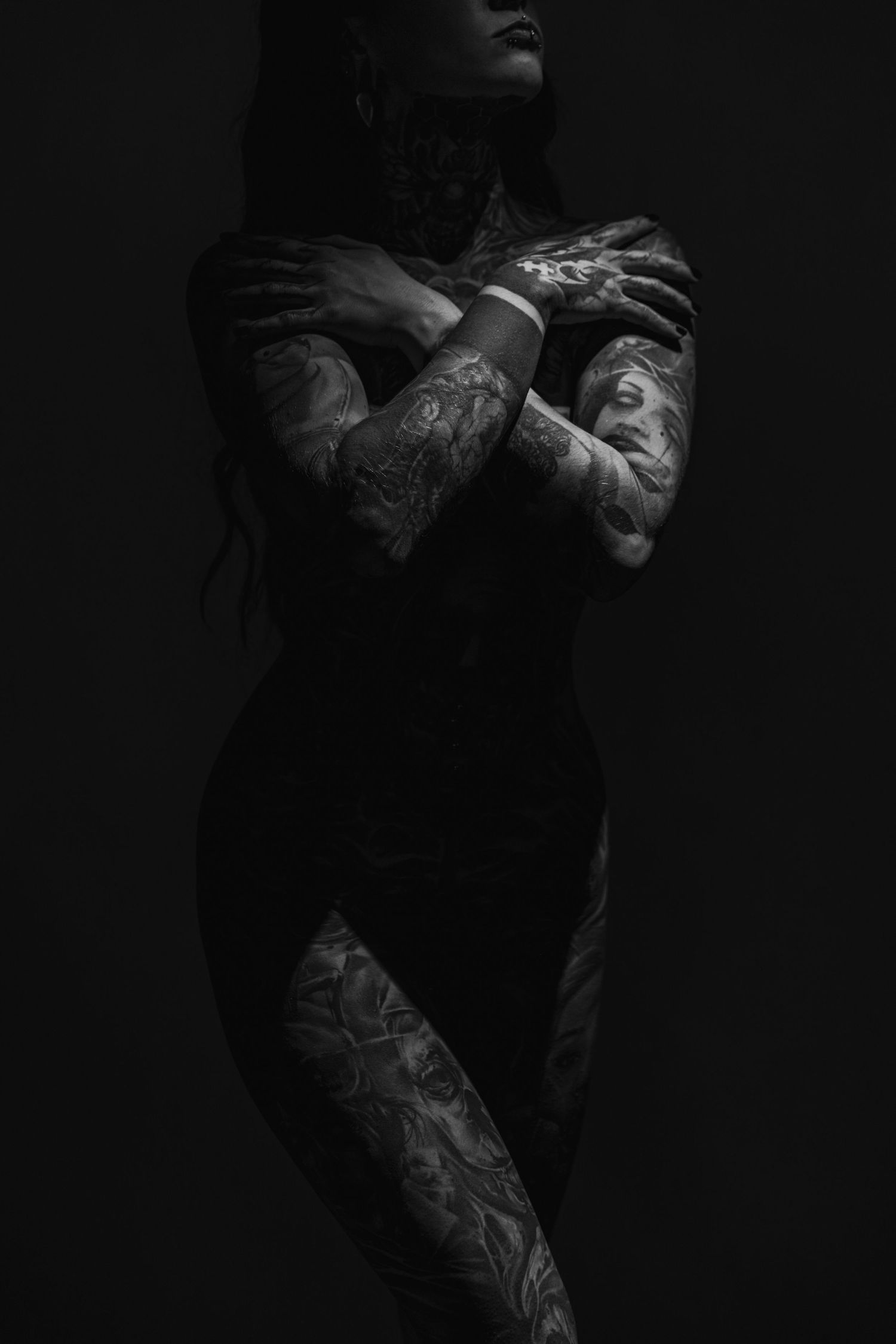 Moody low-key portrait highlighting tattooed arms against black background.