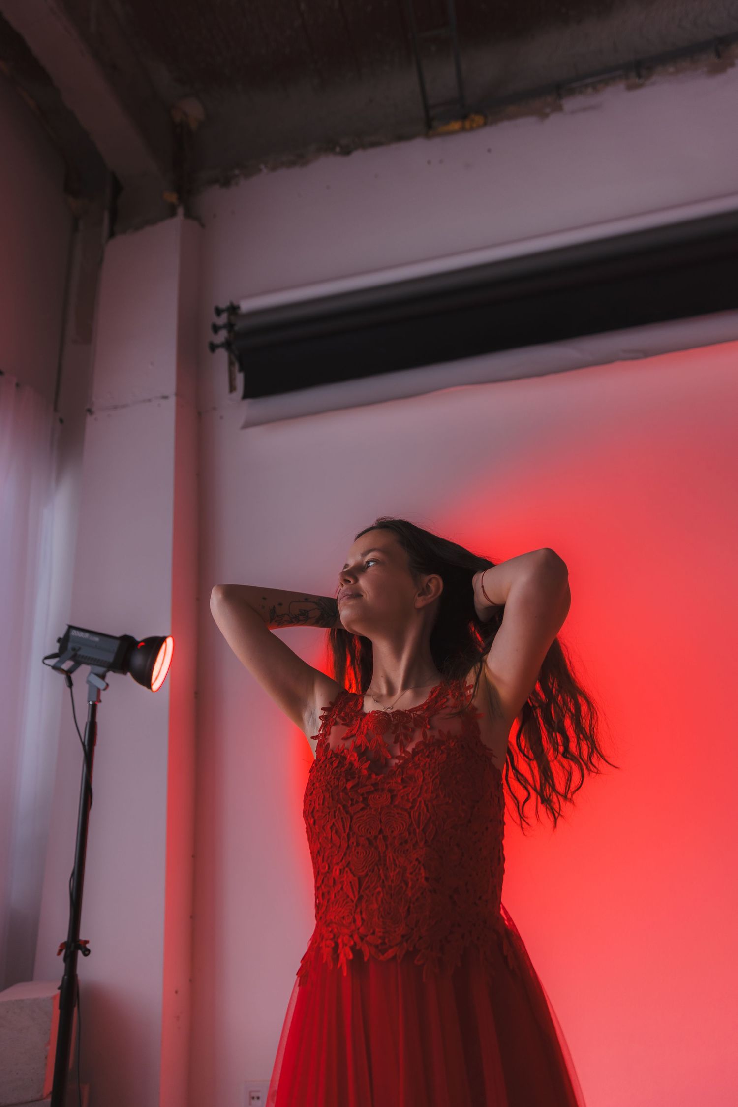 Model in red dress poses against pink-lit studio wall with visible lighting equipment.