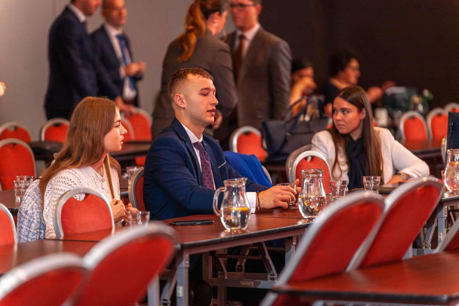 Conference attendees seated at tables with red chairs during business event.