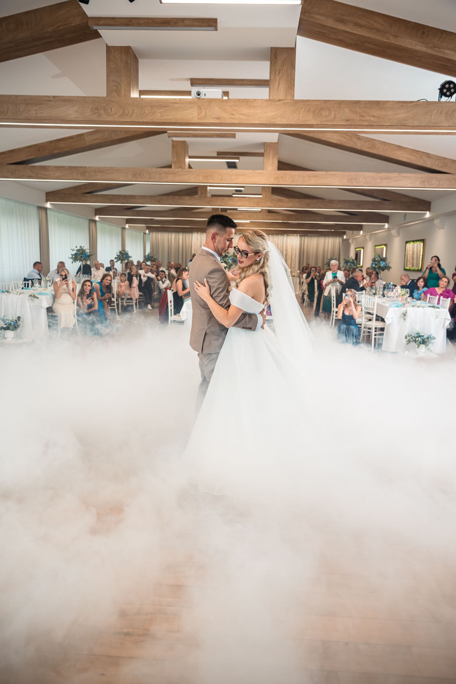 A couple's first dance surrounded by fog effects in a rustic reception venue with wooden beams.