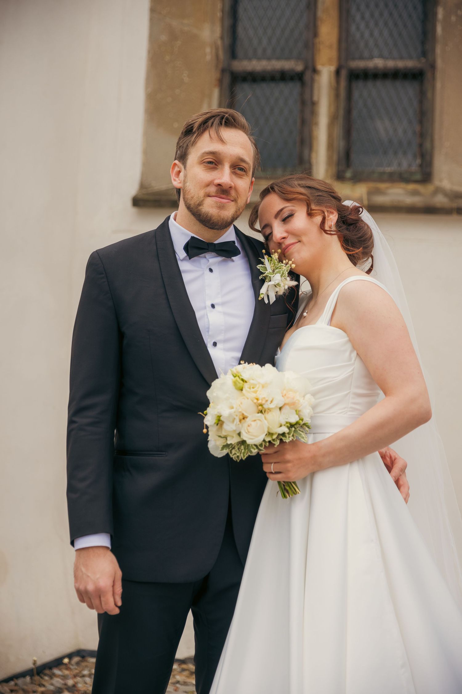 Wedding couple in formal attire poses against neutral wall with vintage windows.