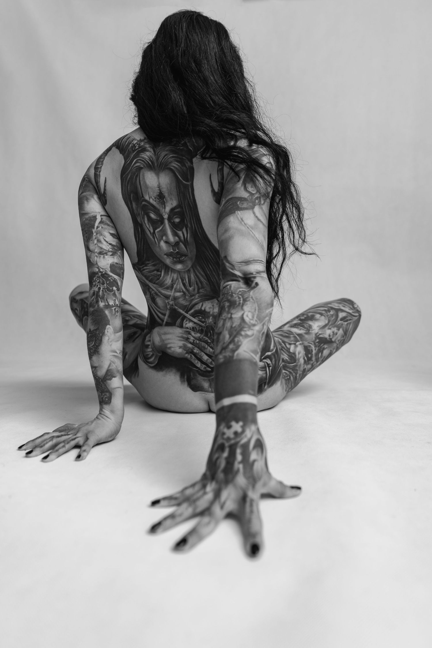 Dramatic monochrome photo of tattooed figure in artistic pose on white backdrop.