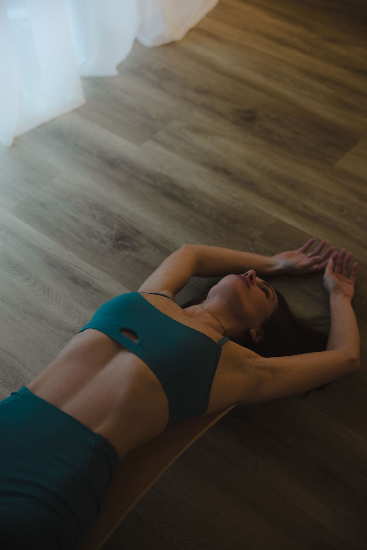 Person lying on wooden floor in teal athletic wear demonstrating relaxation pose.